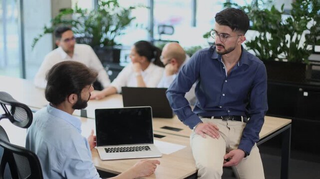 A man in a blue shirt sits at a desk with a laptop and a black laptop
