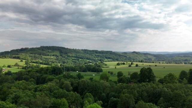 Aerial view of green rolling hills with forest meadows and farmland under a cloudy sky in summer