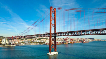 Lisbon's iconic 25 de Abril bridge with its distinctive red towers and suspension cables crosses the Tagus river, connecting the city during a bright, sunny day © Dark Artistic