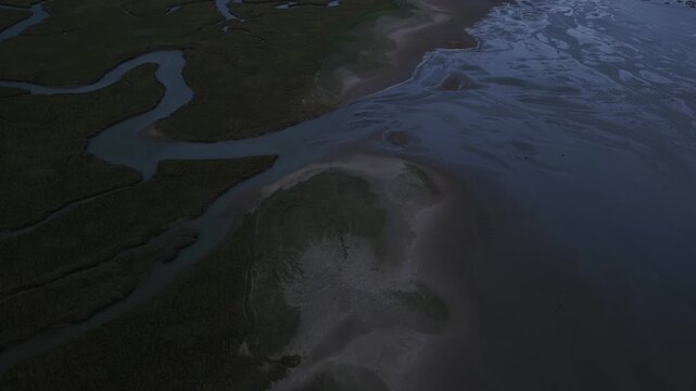 High angle aerial perspective flying over a serene coastal landscape, capturing the intricate patterns of a winding river delta as it merges with the sea during the twilight hours