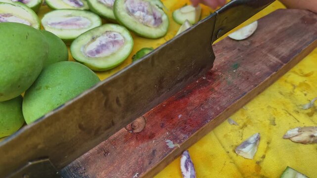 Close view of hands using a traditional Indian mango cutter to slice raw green mango into multiple small pieces for homemade pickle preparation on a yellow surface