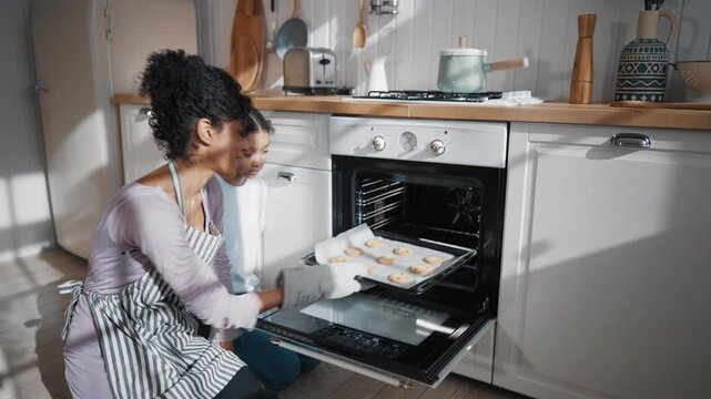 African american woman and her teenage daughter using electric oven in kitchen. Joyful mother and girl baking tasty sweets for family fest diner in weekend, modern home appliance for cook and bake