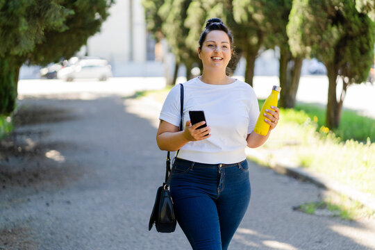 Smiling curvy young woman with mobile phone and bottle in a public park