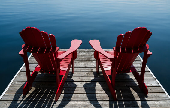 Close up of two red adirondack chairs on a wooden dock facing lake.