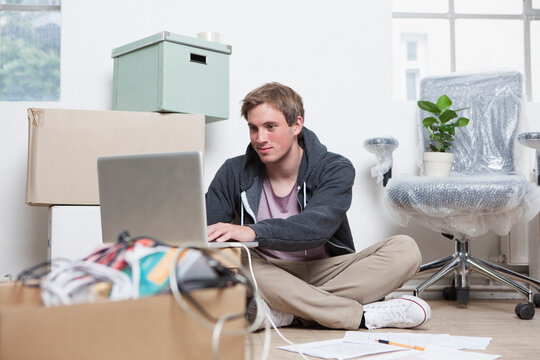 Portrait of young man sitting on ground between cardboard boxes in an office using his notebook