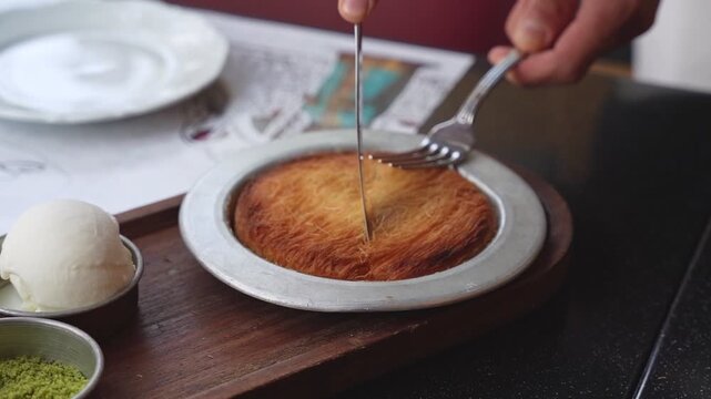 Close-up of a person cutting into a warm, golden Kunafa dessert with a knife and fork. Served with a side of vanilla ice cream and pistachios in a restaurant setting. Bright daylight.