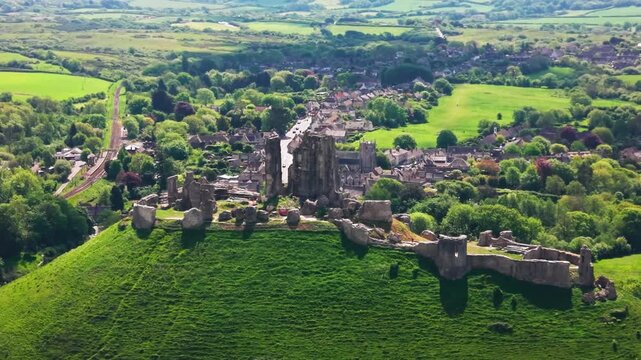 Aerial drone footage of Corfe Castle ruins standing on a green hill above the village of Corfe Castle in Dorset, England. The shot shows the historic medieval castle remains, village streets
