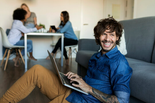 Laughing man sitting on floor using laptop with friends in background