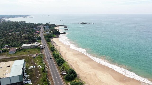 Aerial view of the coastal road running along the sandy beach with turquoise waves and palm trees in Peraliya, Southern Province, Sri Lanka.