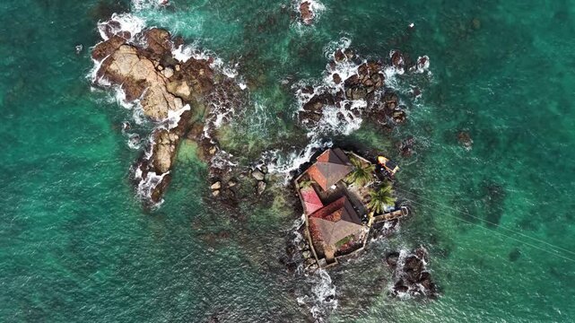 Aerial view of Seenigama Muhudu Dewalaya temple situated on a small rocky island with turquoise waves crashing against the shore Peraliya, Sri Lanka.