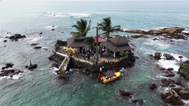 Aerial view of Seenigama Muhudu Dewalaya temple on a rocky island with palm trees and a yellow boat with people in Peraliya, Sri Lanka.