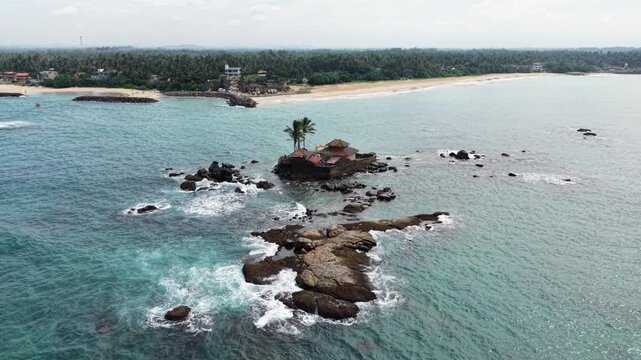Aerial view of Seenigama Muhudu Dewalaya temple on a rocky island with waves crashing against the shore in Peraliya, Sri Lanka.
