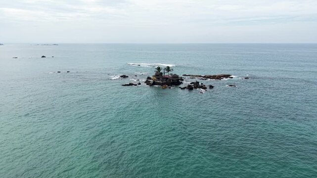 Aerial view of Seenigama Muhudu Dewalaya temple on a small rocky island with palm trees surrounded by turquoise ocean water in Peraliya, Sri Lanka.
