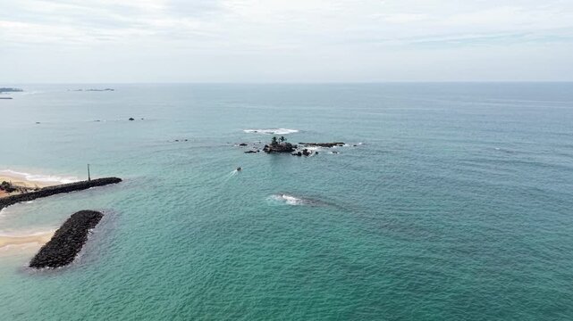Aerial view of Seenigama Muhudu Dewalaya temple on a small rocky island surrounded by turquoise ocean water near the coastline in Peraliya, Sri Lanka.