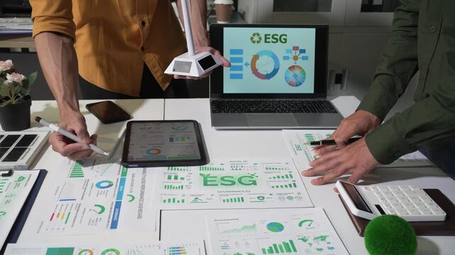 Business team reviewing esg charts and sustainability reports on a desk with laptop, tablet, and calculator during an office meeting on green strategy and corporate planning.