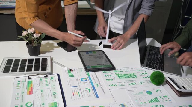 Team reviewing esg reports and renewable energy plans at a desk with wind turbine model, solar panel, tablet and laptop in a modern office.
