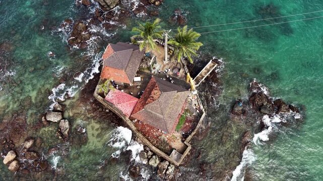 Aerial view of Seenigama Muhudu Dewalaya temple on a rocky island with palm trees surrounded by turquoise ocean waves in Peraliya, Sri Lanka.