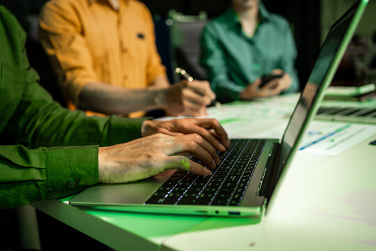 Close view of hands typing on a laptop during a team meeting at night, with coworkers taking notes and using a phone in the background under green office light.