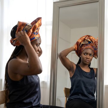 Woman tying vibrant gele headwrap in front of mirror reflecting beauty routine with braids and colorful fabric