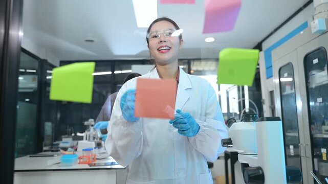 Two female Asian scientists in lab coats discussing research data and planning experiments with sticky notes on a glass board in the laboratory.