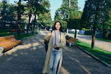 Fototapeta premium Woman in a long beige coat walks along a shaded park path, carrying a camera over her shoulder, casual attire, confident stride amid trees, benches, and a sunny urban park setting