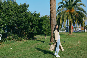 Fototapeta premium Woman in a long beige coat, jeans and sneakers strolls through a sunny park with palm trees, green grass and a relaxed mood, showcasing outdoor fashion