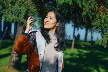 Fototapeta premium Woman in a brown shearling jacket stands outdoors among green trees, sunlight filters through leaves, casual fashion, relaxed pose, cheerful mood, natural park setting, candid lifestyle photograph