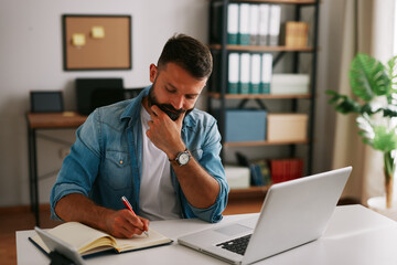 Obraz premium Bearded businessman thinking and taking notes while reviewing data and making decisions at his desk