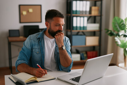 Bearded businessman thinking and taking notes while reviewing data and making decisions at his desk