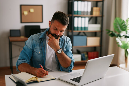 Bearded businessman thinking and taking notes while reviewing data and making decisions at his desk