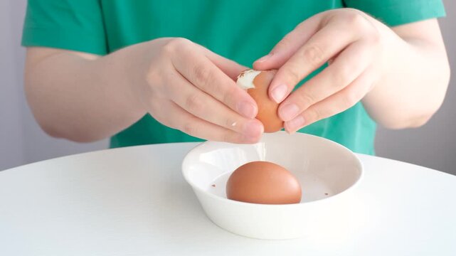 Close-up of female hands peeling the shells of boiled eggs against a white table.