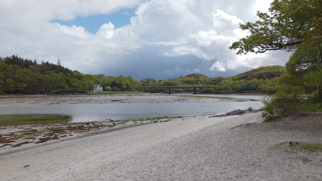 Drone flight pushing slowly over the pristine Silver Sands of Morar towards the Morar Bridge, framed by rolling highland hills and a dynamic sky.