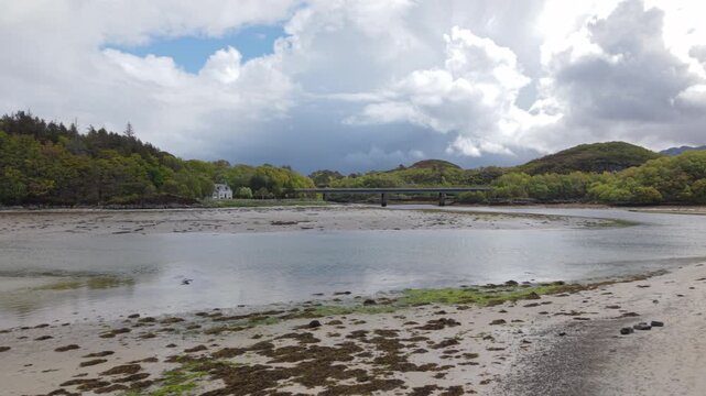 Sideways drone tracking shot across Morar Beach showing the textured tidal sands, with Morar Bridge and the Highland landscape under a dramatic cloudscape.
