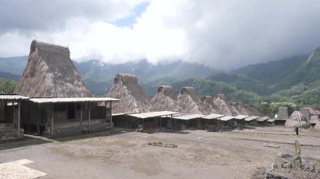 Panoramic view of Gurusina traditional village near Bajawa, Flores, Indonesia, featuring rows of iconic thatched houses beneath mist-covered mountains and lush tropical forest