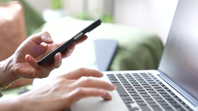 Close up hand holding smartphone while using laptop in modern workspace. Mobile communication, remote work, online business, digital connectivity and multitasking productivity concept.work from home