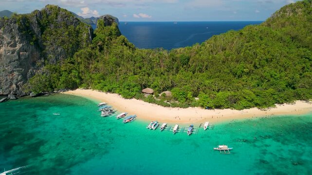 Aerial orbit of Helicopter Island beach with row of tour boats along turquoise reef white sand and lush limestone cliffs in Bacuit Bay of El Nido Palawan Philippines