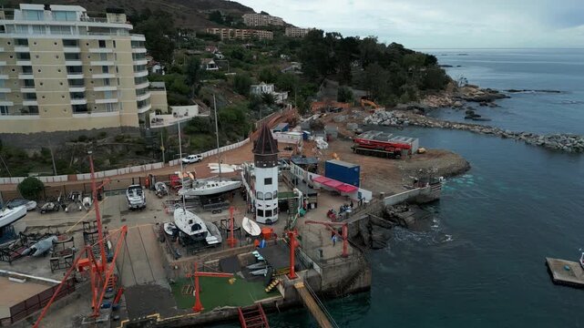 Slow revolving shot over active boatyard with cranes and dry-docked vessels beside coastal apartment tower and rocky headland. Papudo, Chile