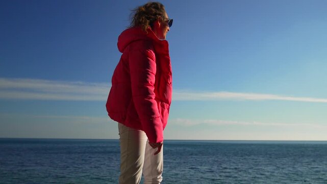 Woman sea jacket woman wearing a bright pink puffer coat stands at the seashore looking at the horizon on a sunny day