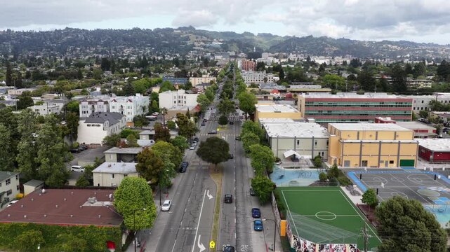 Tree-lined urban boulevard with traffic, school sports field, mixed residential and commercial buildings stretching toward densely forested hills under overcast sky. California, USA. Aerial wide shot.