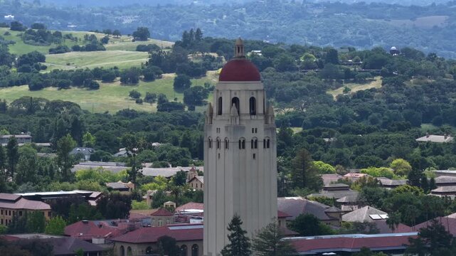 Hoover Tower with red dome rising above red tile campus buildings and lush tree canopy with rolling green hills and oak woodland stretching beyond under overcast sky. California, USA. Aerial wide shot