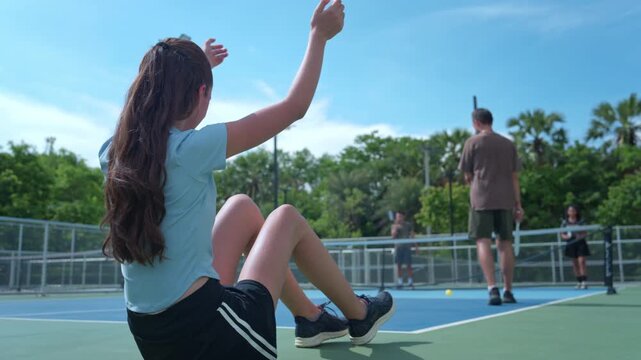 A young female pickleball players sit by the sidelines of an outdoor field, cheering on their friends who are competing in a pickleball game, demonstrating the enjoyment they have playing a recreation