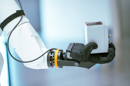 A close-up of a white robotic arm featuring a black fabric-covered end effector as it holds a gray electronic component during a tech demonstration.