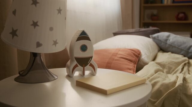 Close-up view of toy rocket and book resting on bedside table in kids bedroom next to unmade bed. Clip serving as perfect visual for family lifestyle branding, parenting ads