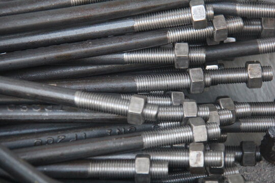 Close-up of steel threaded rods with nuts and bolts stacked in an industrial workshop. Metal fasteners background, construction hardware, engineering and manufacturing concept.