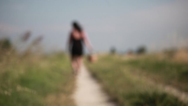 Young woman walking away on rural path carrying basket and picking flowers. Blurred countryside landscape, raw footage, ungraded natural light. Summer lifestyle.