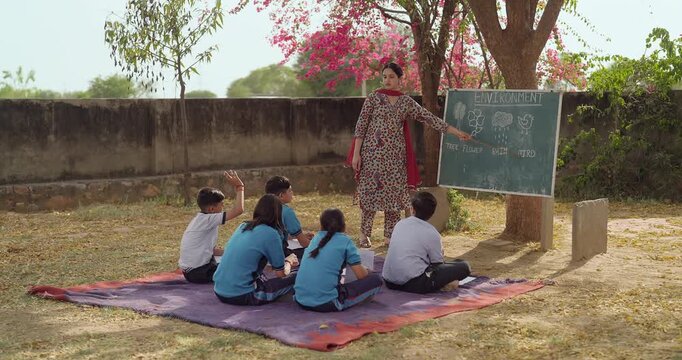 Indian young adult female tutor hold stick pointing words teach to girl boy rural area place. Village group sitting on mat look board talk speak read do study learn clap hands open ground class field