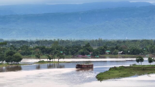 Aerial view of a traditional houseboat cruising through the calm waters of Tanguar Haor with lush greenery and blue hills in Sunamganj District, Sylhet Division, Bangladesh.