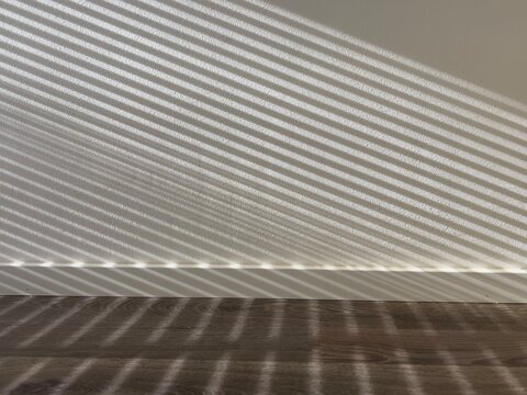 Dramatic parallel shadows from window blinds are cast diagonally across a plain white wall and a dark wooden floor, creating a stark, minimalist pattern of light and dark.