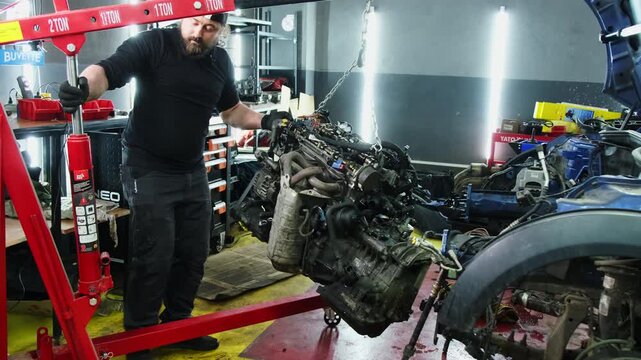 Mechanic moves engine with crane before repair work. Male technician positions large part touching special metal construction with hands