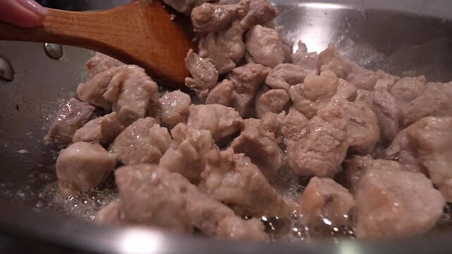 Pork pieces sizzling in a hot stainless steel frying pan while being stirred with a wooden spatula, a delicious and easy meal preparation showing the browning process for a tasty stew slow motion